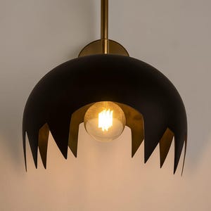 May include: A modern wall sconce with a black, dome-shaped shade and a brass-colored stem and accents. The shade has a jagged, decorative edge, and a visible light bulb is centered within the design. The fixture is mounted on a white wall.