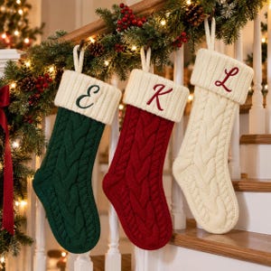 Puede incluir: Tres calcetines navideños de punto trenzado con puño acanalado. El primero es verde oscuro con la letra E bordada, el segundo es rojo con la letra K bordada, y el tercero es color crema con la letra L bordada en color rojo.