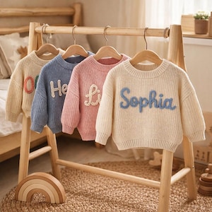 Op de afbeelding: Vier kindertruien in beige, blauw, roze en crème, elk met een naam op de voorkant geborduurd. De truien hangen aan een houten rek. De namen zijn in verschillende kleuren geborduurd.