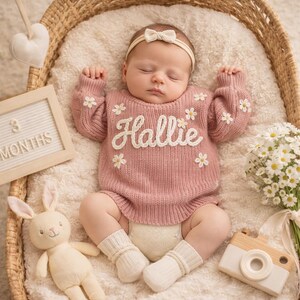 Op de afbeelding: Een slapende baby in een roze gebreide romper met de naam "Hallie" in witte letters en madeliefjesaccenten. De baby ligt in een rieten mand met een zachte deken, omringd door een speelgoedkonijn, een houten camera en een bord met "3 MAANDEN".