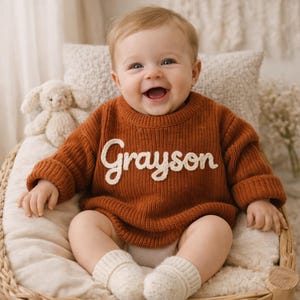 Op de afbeelding: Een lachende baby draagt een roestkleurige gebreide trui met de naam "Grayson" in witte letters. De baby zit in een rieten stoel, draagt witte sokken en heeft een knuffelkonijn in de buurt. De trui heeft lange mouwen.