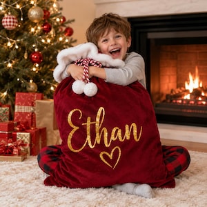 Pode incluir: Um saco de Pai Natal em veludo vermelho profundo com rebordo em pelo sintético branco no topo e cordões torcidos em vermelho e branco com pompons. O nome Ethan está escrito em glitter dourado sobre um coração de glitter dourado.
