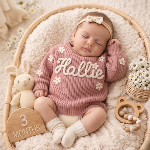 Op de afbeelding: Een slapende baby in een roze gebreide trui met de naam "Hallie" in witte letters en madeliefjesaccenten. De baby ligt in een rieten mand met een pluchen konijn, een houten "3 MAANDEN" bord, een houten bijtring en een klein mandje met madeliefjes.