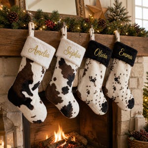 Puede incluir: Cuatro calcetines navideños de felpa con estampado de vaca. Dos calcetines tienen puños blancos bordados con los nombres dorados Amelia y Sophia, y dos tienen puños negros bordados con los nombres dorados Olivia y Emma.