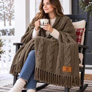 May include: A chunky olive green knit throw blanket featuring a vertical cable knit pattern and long fringe trim along the bottom edge. A rectangular tan leather patch with the text The Woolloom is stitched onto the lower right corner.