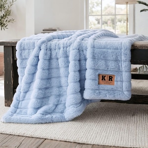 May include: A light blue faux fur throw blanket with a textured, ribbed pattern. A rectangular tan leather patch is sewn onto the corner, featuring the black printed text K R, the names Katie and Ryan separated by a heart, and the date 12.10.2019.
