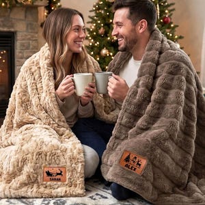 May include: Two plush faux fur blankets, one in cream and one in taupe, each featuring a rectangular leather patch. The cream blanket patch displays a sleigh graphic with the name SARAH, while the taupe blanket patch shows a snowman and the name ALEX.