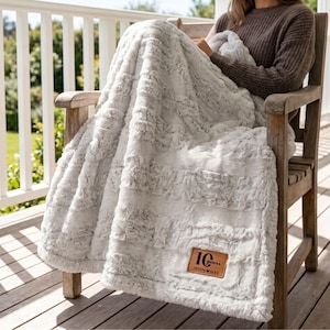 May include: A soft, light gray faux fur throw blanket with a textured, swirling pattern. A rectangular tan leather patch is stitched onto the corner, featuring the embossed text 10 years, a heart symbol, and the names Olivia and Alex.