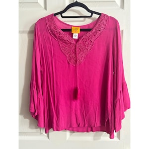 May include: A bright pink blouse with long sleeves and bell cuffs. The neckline features intricate lace and bead detailing, with a tassel. The blouse is hanging on a black hanger against a white door.