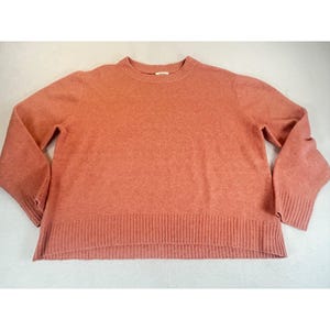 May include: A solid terracotta-colored long-sleeve sweater featuring a crew neckline and a ribbed knit texture on the collar, cuffs, and hem. The garment has a relaxed fit with a slightly longer back hemline and a small white brand label at the neck.