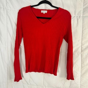 May include: A bright red, long-sleeved sweater with a V-neck. The sweater has a ribbed texture and a small label near the neckline. The sleeves have a slight flare at the cuffs. The sweater is hanging on a black hanger.