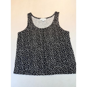 May include: A black sleeveless tank top featuring a repeating white speckled pattern throughout the fabric. The garment has a scoop neckline and a central vertical seam running down the front. A white label is visible at the inner neckline.