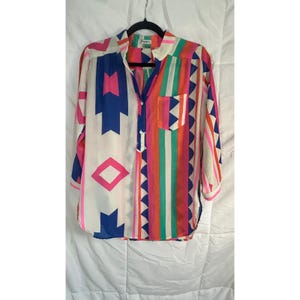 May include: A long-sleeve button-up shirt with a colorful geometric pattern. The shirt features a mix of blue, pink, orange, and white shapes and stripes. It has a single pocket and a collared neckline.