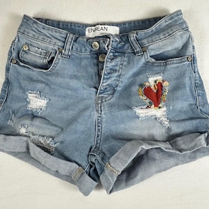 May include: Light blue denim shorts with distressed details and a rolled hem. Features a red heart patch with floral embroidery. The shorts have a button closure and pockets. The brand name "EN EAN" is visible on the label.