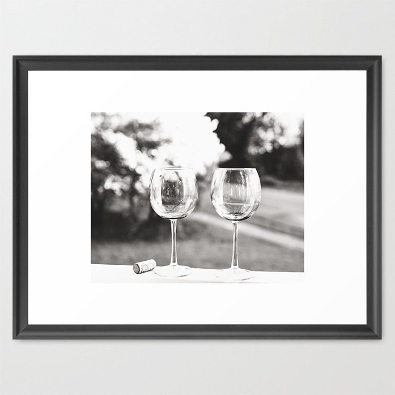 May include: Two empty wine glasses sit on a white surface with a cork in the foreground. The glasses are clear and have a round bowl and a long stem. The background is out of focus and shows a blurry image of trees and foliage.