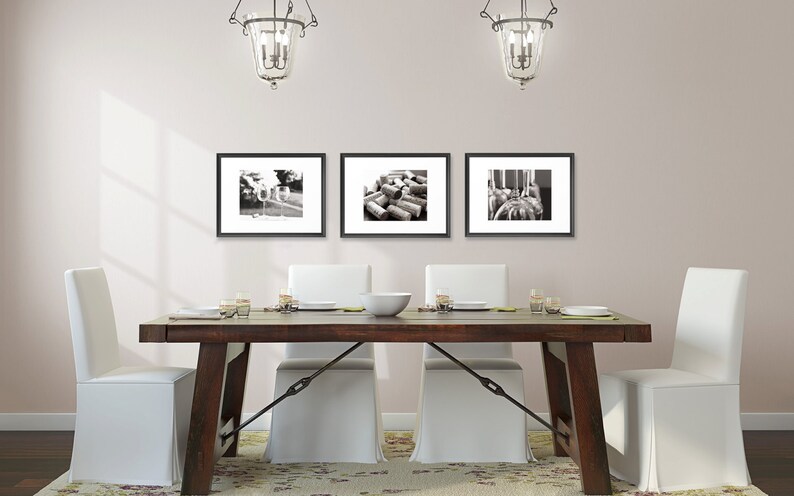 May include: A dining room scene featuring a dark wood table, white chairs, and three framed black and white photographs. The photographs depict wine-related imagery. Two ornate light fixtures hang above the table, and the room has a neutral color palette.