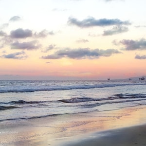 May include: A scenic sunset over the ocean with a sandy beach in the foreground. The sky is a mix of pink, orange, and purple hues with fluffy white clouds. The ocean is a deep blue with white waves crashing on the shore.
