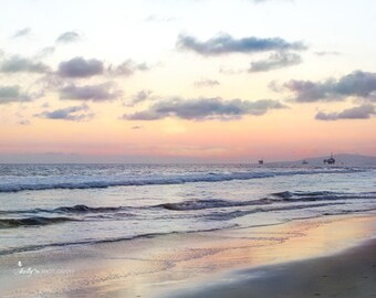 May include: A scenic sunset over the ocean with a sandy beach in the foreground. The sky is a mix of pink, orange, and purple hues with fluffy white clouds. The ocean is a deep blue with white waves crashing on the shore.