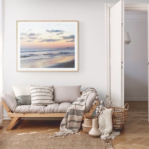 May include: A living room interior with a light gray couch, a wooden coffee table, and a framed photograph of a beach scene with a sunset sky. The couch has a white and gray striped throw pillow and a white throw blanket with black stripes. There is a white vase with a dried flower arrangement on the coffee table.