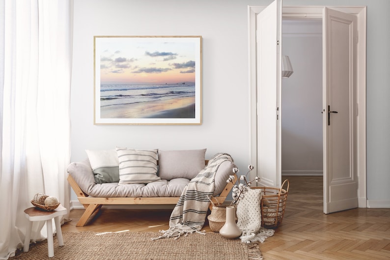 May include: A living room interior with a light gray couch, a wooden coffee table, and a framed photograph of a beach scene with a sunset sky. The couch has a white and gray striped throw pillow and a white throw blanket with black stripes. There is a white vase with a dried flower arrangement on the coffee table.