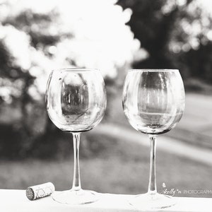 May include: Two empty wine glasses on a white surface with a blurred background of trees and a cork on the left side of the image.