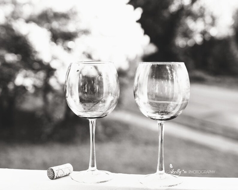 May include: Two empty wine glasses on a white surface with a blurred background of trees and a cork on the left side of the image.