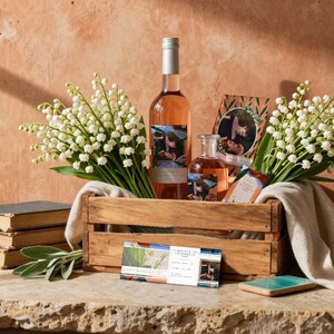 May include: A wooden crate filled with a bottle of rosé wine, a smaller bottle, and a framed photo. White lily of the valley flowers and a stack of books are also present. A business card and a small blue tile complete the arrangement.