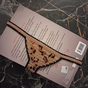 May include: A handmade crochet bookmark with a leopard print design. The bookmark is tan with black accents and is lying on top of a book. The bookmark is made of yarn and has a unique, textured appearance.