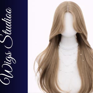 May include: A light brown wig with long, layered hair is displayed on a mannequin head. The wig features a center part and a soft wave. A multi-strand pearl necklace adorns the mannequin. The words "Wigs Studio" are written in white script on a dark blue background.