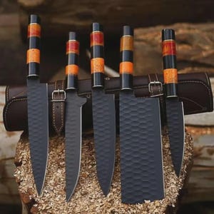 May include: A set of five kitchen knives with matte black hammered blades and cylindrical handles featuring alternating bands of black, orange, and yellow resin. The knives are arranged in front of a brown leather roll case with metal buckles.