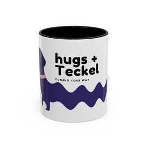 May include: White ceramic mug with a black interior and the words "hugs + Teckel" in black. A purple dachshund illustration and a purple wavy line are on the side. The text "COMING YOUR WAY" is below the main text.