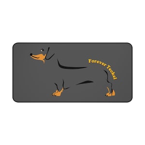 May include: A rectangular mousepad featuring a stylized black dachshund dog with orange accents. The words "Forever Teckel" are written in yellow cursive. The background is a solid dark gray.