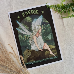 Puede incluir: Una impresión que presenta una hada sentada con largo cabello rubio y delicadas alas azules y blancas. El hada lleva un vestido gris claro y está situada sobre un fondo oscuro con la palabra "FAERIE" en verde.
