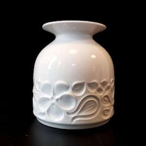 May include: A white ceramic vase with a bulbous body and a flared rim. The vase features a raised floral pattern around the base, with detailed flower and leaf designs. The background is solid black, highlighting the vase's shape and texture.