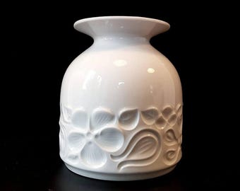 May include: A white ceramic vase with a rounded body and a flared rim. The vase has a raised floral pattern around the base, with detailed flower and leaf designs. The background is solid black, highlighting the vase's shape and texture.