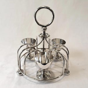 Op de afbeelding: Een zilverkleurige eierhouder met een cirkelvormige handgreep en een ronde basis. Het heeft vijf eierdopjes, elk met een voet. De houder is ontworpen met gebogen metalen steunen en een centrale steel.