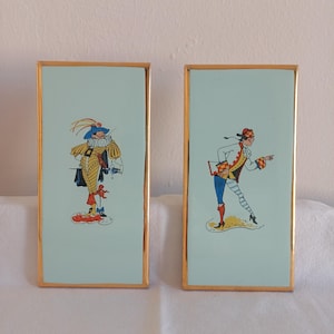Può includere: Due opere d'arte rettangolari incorniciate. Ognuna presenta uno sfondo azzurro con un bordo dorato. Una raffigura un personaggio dei cartoni animati in un abito giallo e nero con un cappello blu. L'altra mostra una figura in un abito bianco, rosso e blu.