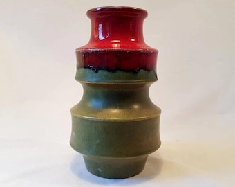 May include: A ceramic vase with a layered design. The top is a glossy red, transitioning to a dark red band, then a green section, and finally a larger olive green base. The vase has a unique, textured appearance.