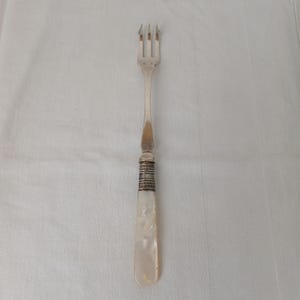 Peut inclure: Une fourchette en argent à trois dents et un manche décoratif. Le manche est orné d'une poignée en nacre avec des bandes argentées. La fourchette est conçue pour servir ou manger, et est probablement vintage ou antique.