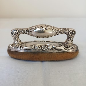 Op de afbeelding: Een antiek zilverkleurig naai-instrument met een handvat en een ronde, bruine kussenbasis. Het handvat heeft sierlijke bloemendetails. Het kussen is lichtbruin van kleur. Het gereedschap wordt waarschijnlijk gebruikt voor het persen van naden.