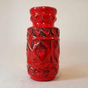 Może przedstawiać: Żywy, czerwony wazon ceramiczny z teksturowanym, geometrycznym wzorem. Wazon ma cylindryczny kształt z rozszerzaną górą i podstawą. Projekt obejmuje podniesione kształty rombów i okręgów z ciemnymi akcentami. Wazon ma około 25 cm wysokości.