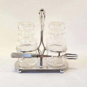 Könnte beinhalten: Ein Vintage-Gewürzset aus Silber. Es besteht aus zwei Gläsern mit Diamantschliff und passenden Deckeln. Das Set enthält eine kleine Gabel und ein Messer mit Perlmuttgriff, alles auf einem Silbertablett.