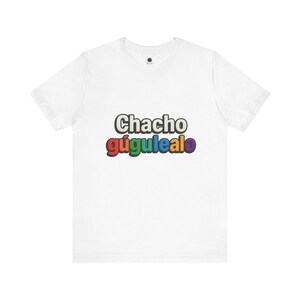 May include: White t-shirt with the text "Chacho gúgulealo" in a colorful, outlined font. The text is in a playful style, with the word "Chacho" in a larger font size than "gúgulealo". The shirt has a crew neck and short sleeves.