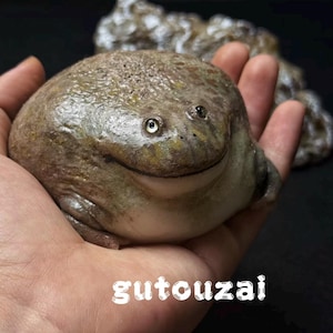 May include: A round, textured, brown and green frog figurine with a smooth, light-colored belly and a wide, smiling mouth. The frog has small, dark eyes and a bumpy, mottled skin surface. The text gutouzai is printed in white at the bottom.
