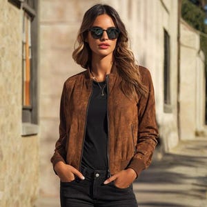 May include: A brown suede bomber jacket with a zip fastening. The jacket features ribbed cuffs and hem. The person is wearing black sunglasses, a black top, and black jeans. The background is a stone building.