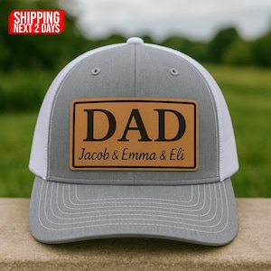 Może przedstawiać: Szara i biała czapka typu trucker z brązową skórzaną naszywką z napisem "DAD" nad imionami "Jacob & Emma & Eli". Czapka ma zakrzywiony daszek i siatkową tylną część.