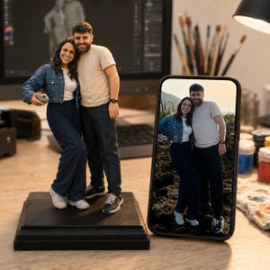 May include: A 3D printed figurine of a couple standing on a black square base, positioned next to a smartphone displaying a photograph of the same couple. The figurine depicts a woman in a denim jacket and dark pants and a man in a beige t-shirt and dark pants.