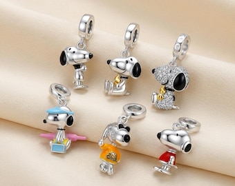May include: Six silver-toned metal charms featuring the character Snoopy in various poses and outfits. One charm is covered in clear crystals, another wears a blue cap with a pink accessory, one wears an orange shirt with the word COOL, and one wears red.