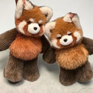 May include: Two plush red panda stuffed animals. The plush toys have brown and orange fur with white accents around the eyes and ears. The stuffed animals are standing next to each other.