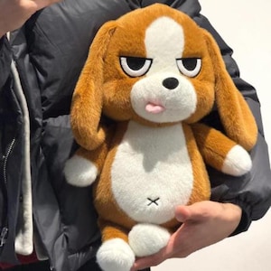 Könnte beinhalten: Ein Plüschhundespielzeug mit braunem und weißem Fell. Der Hund hat einen grimmigen Gesichtsausdruck mit schwarzen Augenbrauen, einer rosa Zunge und einem schwarzen 'X' auf der Brust. Das Spielzeug hat große, schlaffe Ohren und weiße Pfoten.